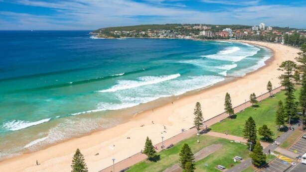 A coastal beach with a paved walkway, a parking area, and a view of the ocean and distant buildings. - Comfy 2-Bed Apartment with A/C throughout (Manly)