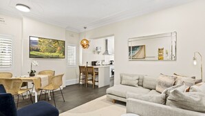 A modern living room with a sofa, dining table, and chairs. There is a kitchen area with a dining island and chairs. A large mirror and artwork are on the walls. - Comfy 2-Bed Apartment with A/C throughout (Manly)