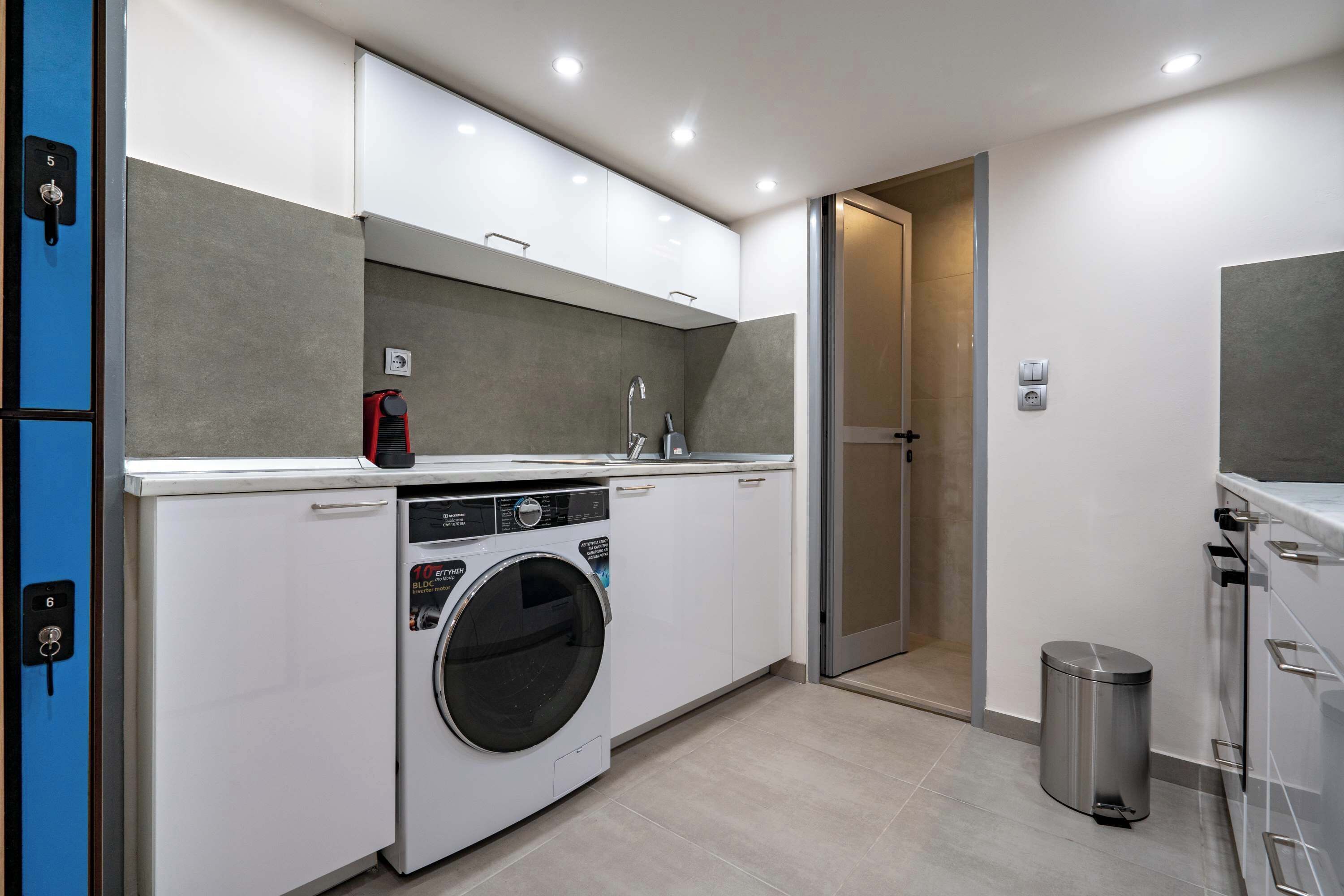 A compact laundry room with a front-loading washing machine, a small sink, and a trash can.