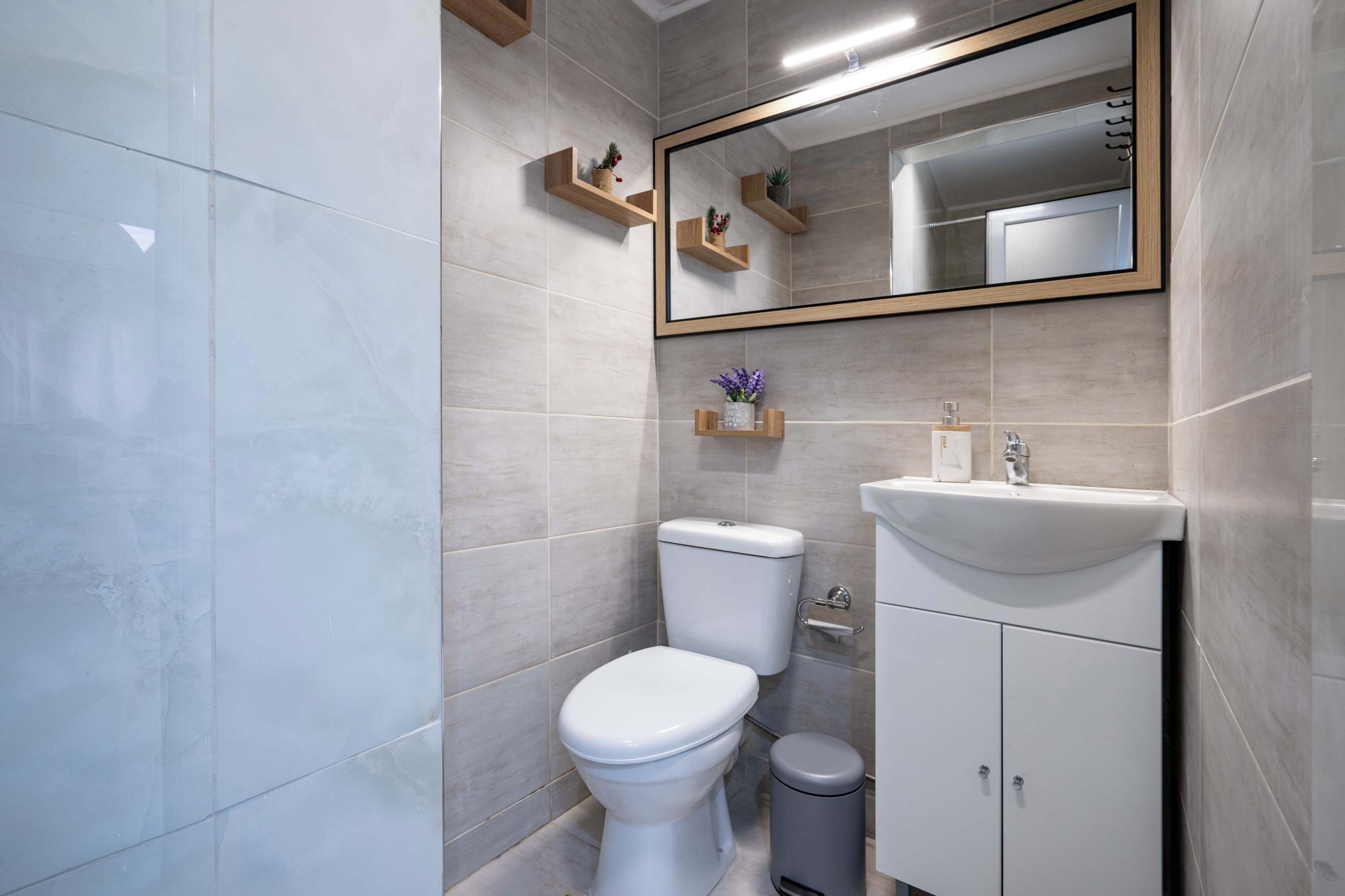 A modern bathroom with a toilet, sink, and mirror. There are wooden shelves with plants and a trash bin.