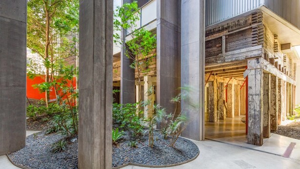 Un bâtiment moderne avec des piliers en béton, un petit espace jardin avec des plantes et une structure en bois dotée d’un escalier.