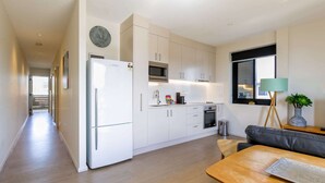 A modern kitchen with white cabinets, a refrigerator, microwave, and oven. There is a dining area with a wooden table and a black leather couch. - Comfy 2-Bedroom Apartment with a Clyde River View (North Batemans Bay)