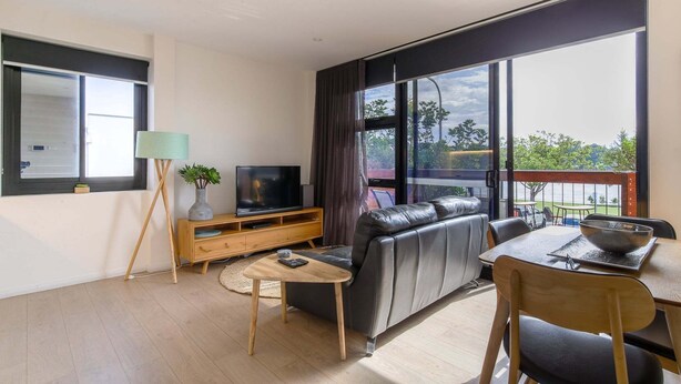A modern living room with a leather sofa, a TV on a wooden stand, a dining table with chairs, and large windows offering a view of the outdoors. - Comfy 2-Bedroom Apartment with a Clyde River View (North Batemans Bay)
