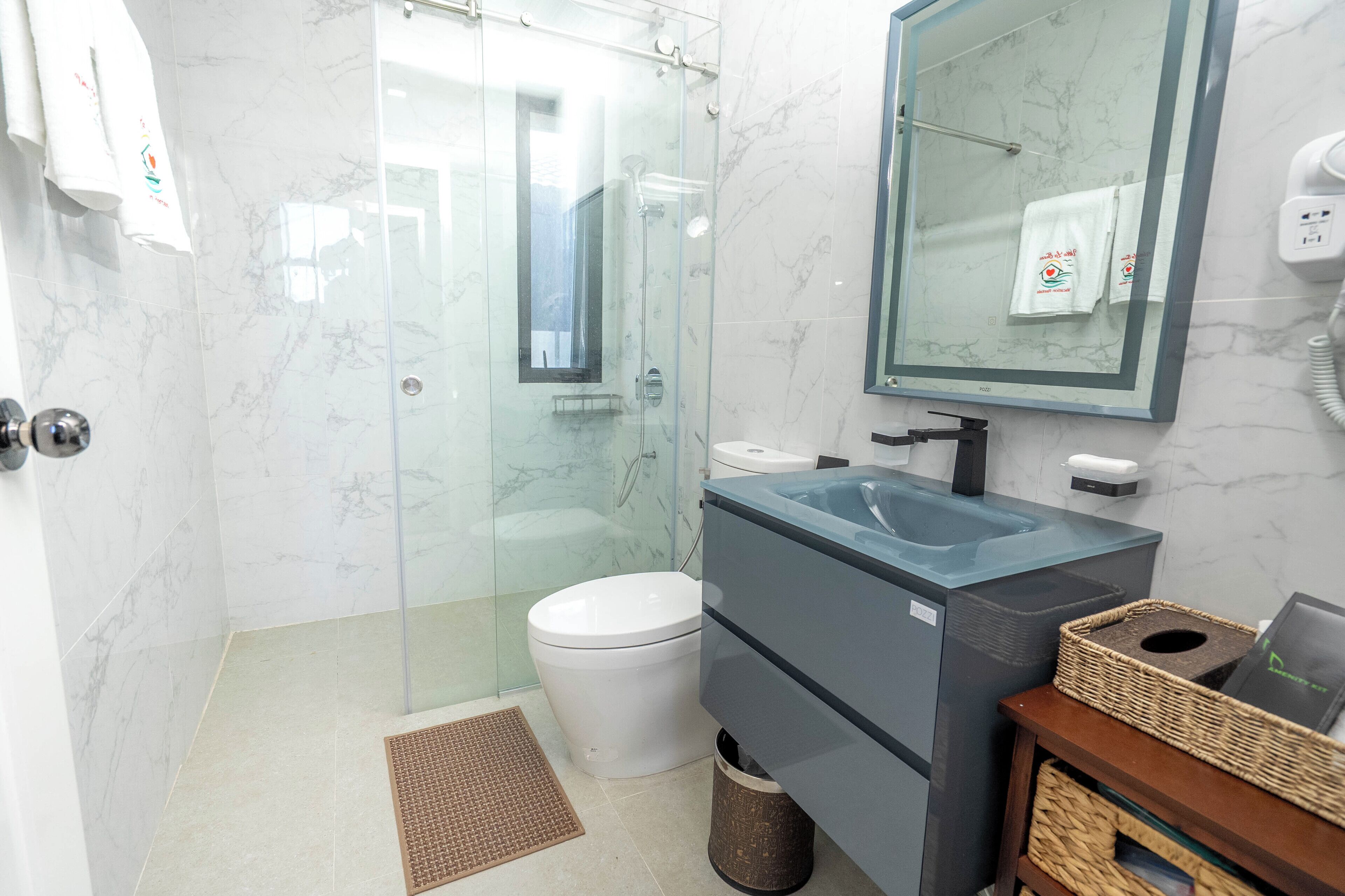 Un baño moderno con ducha envuelta en vidrio, inodoro blanco, lavamanos con encimera de mármol y espejo sobre el lavamanos.