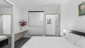 Une chambre d’hôtel moderne avec un grand lit, une table de chevet avec une lampe, un petit coin salon agrémenté d’un vase de fleurs et une porte donnant sur une autre pièce.