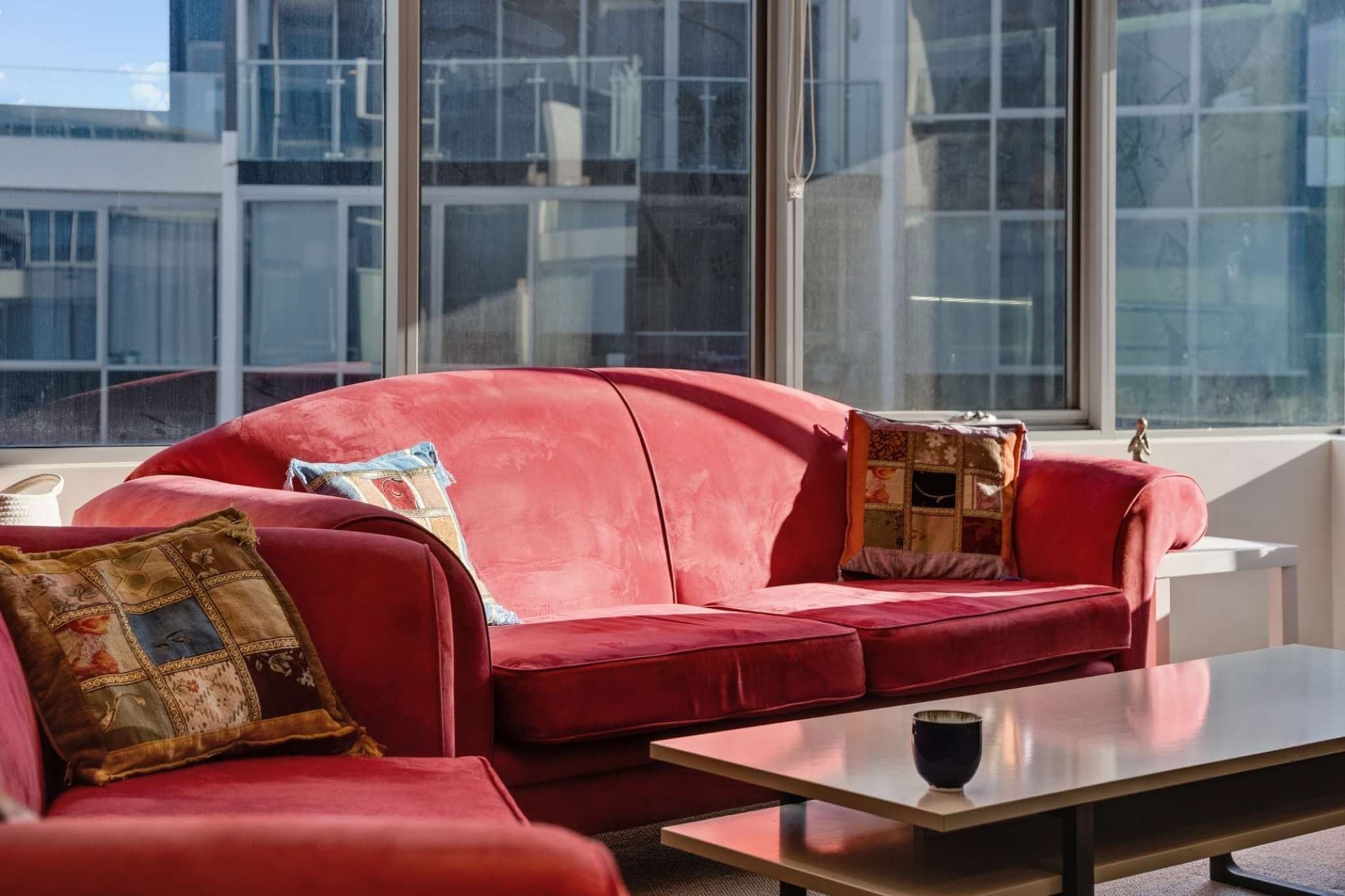 Un sofá rojo con cojines estampados, una mesita con una taza, y grandes ventanales con vistas a un edificio.