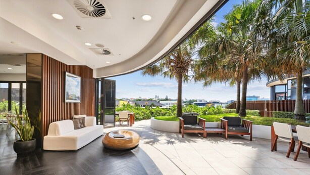 A modern interior with a curved ceiling, a white sofa, a wooden coffee table, and a view of palm trees and a cityscape. - Chic 1-Bed with Parking, Pool and Gym (Fortitude Valley)