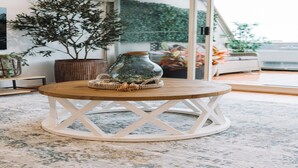 Een ronde houten tafel met een glazen vaas op een geweven onderzetter, een potplant en uitzicht op een balkon met planten en een balustrade.