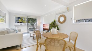 A modern living room with a white sofa, wicker chairs, a wooden dining table, and a TV. - Ground Floor 2-Bed Mermaid Beach Pad with Parking (Mermaid Beach)