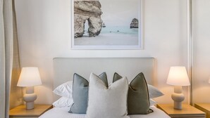Een net opgemaakt bed met kussens en twee bedlampjes, een lijst met een strandafbeelding boven het bed, en een houten hoofdbord.