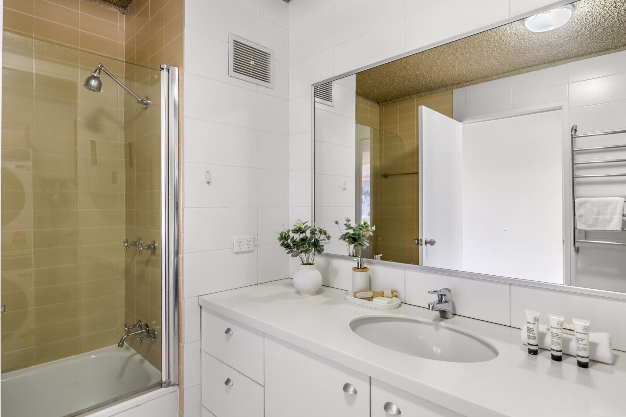 A modern bathroom with a glass-enclosed shower, a large mirror, and a white vanity with a sink and toiletries.