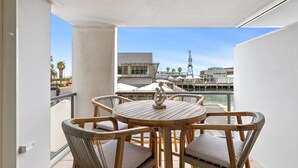Un balcón con una mesa redonda de madera y sillas de mimbre, con vista a un puerto deportivo con barcos y edificios.