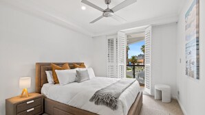 Un dormitorio con cama, mesita de noche, lámpara, ventilador de techo, ventana abierta y vistas al exterior.