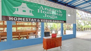 Een groene banner met de tekst 'Anggrek Homestay Jogja', houten banken, een rode kast en een betonnen vloer.