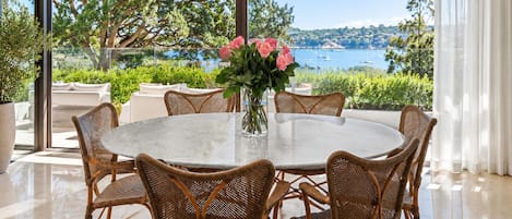 A round dining table with wicker chairs, a vase with flowers, and a view of a lake and trees.