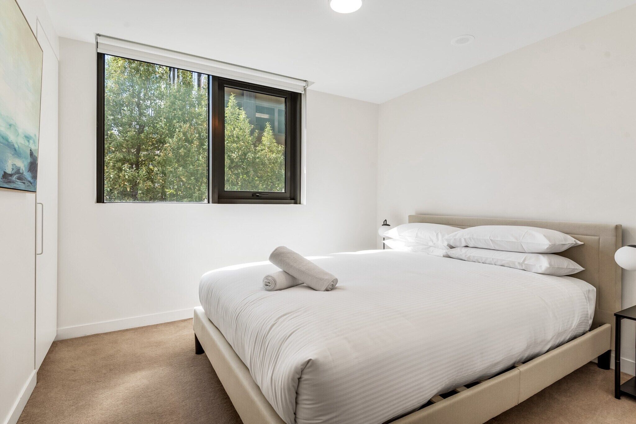A bedroom with a large window, a bed with white linens, and a nightstand.
