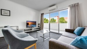 A modern living room with a flat-screen TV, grey and brown sofas, a glass coffee table, and a sliding glass door leading to a balcony.