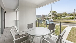 Un balcón con una mesa redonda y sillas, una puerta de cristal y vistas a una carretera y vegetación.