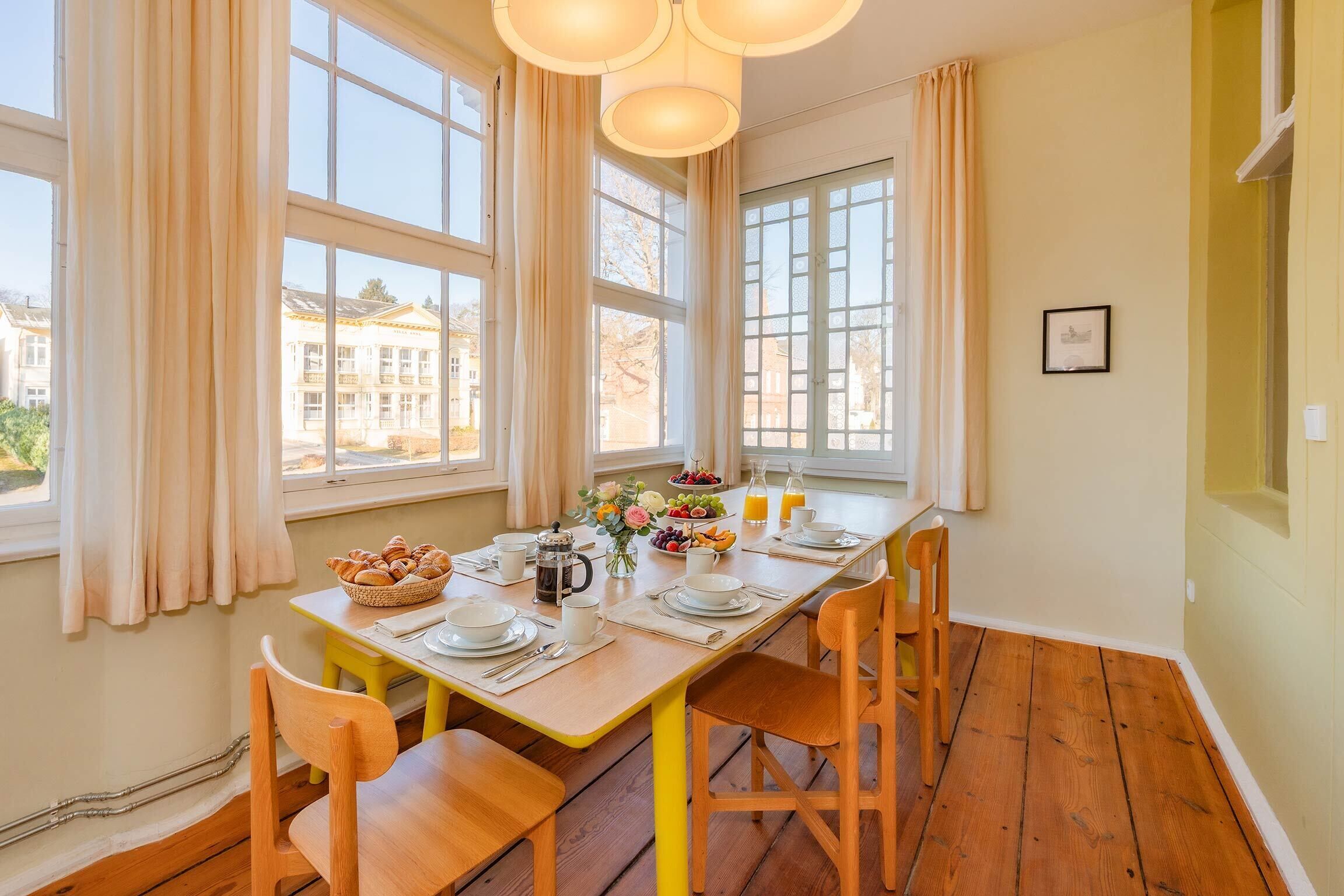 Ein Essbereich mit einem Holztisch, der für ein Frühstück gedeckt ist, großen Fenstern und Blick auf Gebäude im Freien.