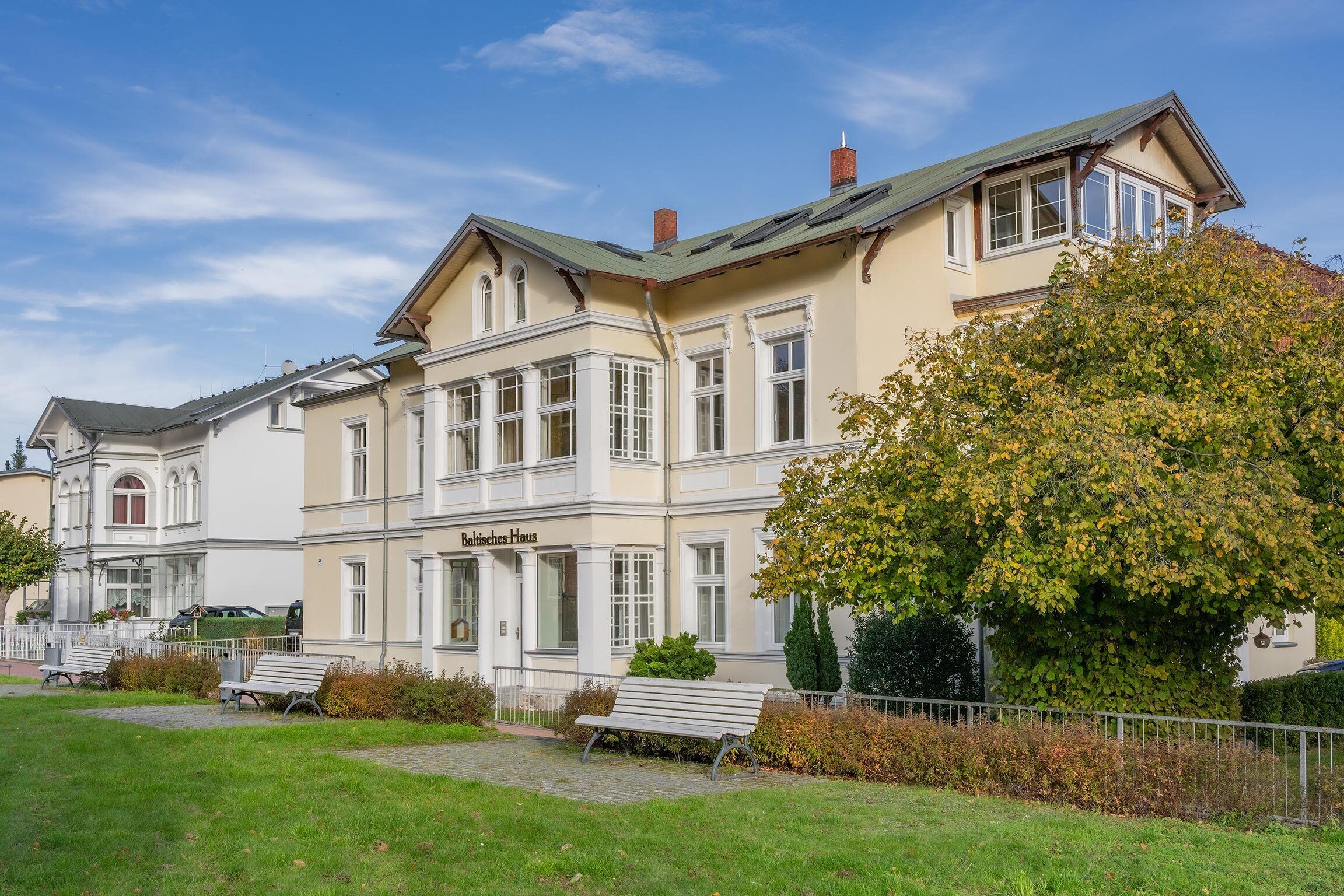 En traditionell byggnad med en framträdande entré, flera fönster och en välskött gräsmatta med bänkar.