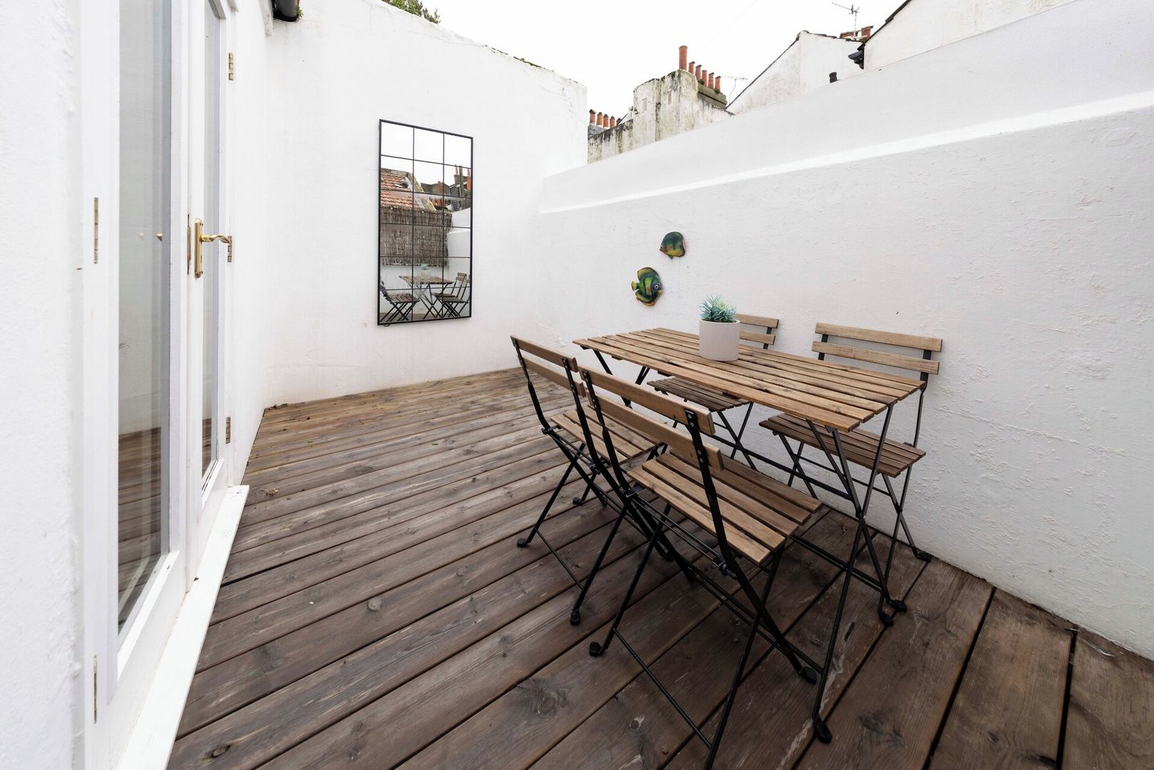 A wooden deck with a table and chairs, a white wall, and a window with a view of buildings.