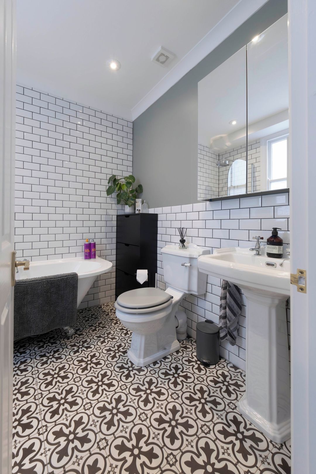 A bathroom with white subway tiles, a black patterned floor, a white toilet, a black cabinet, and a white sink with a mirror above.