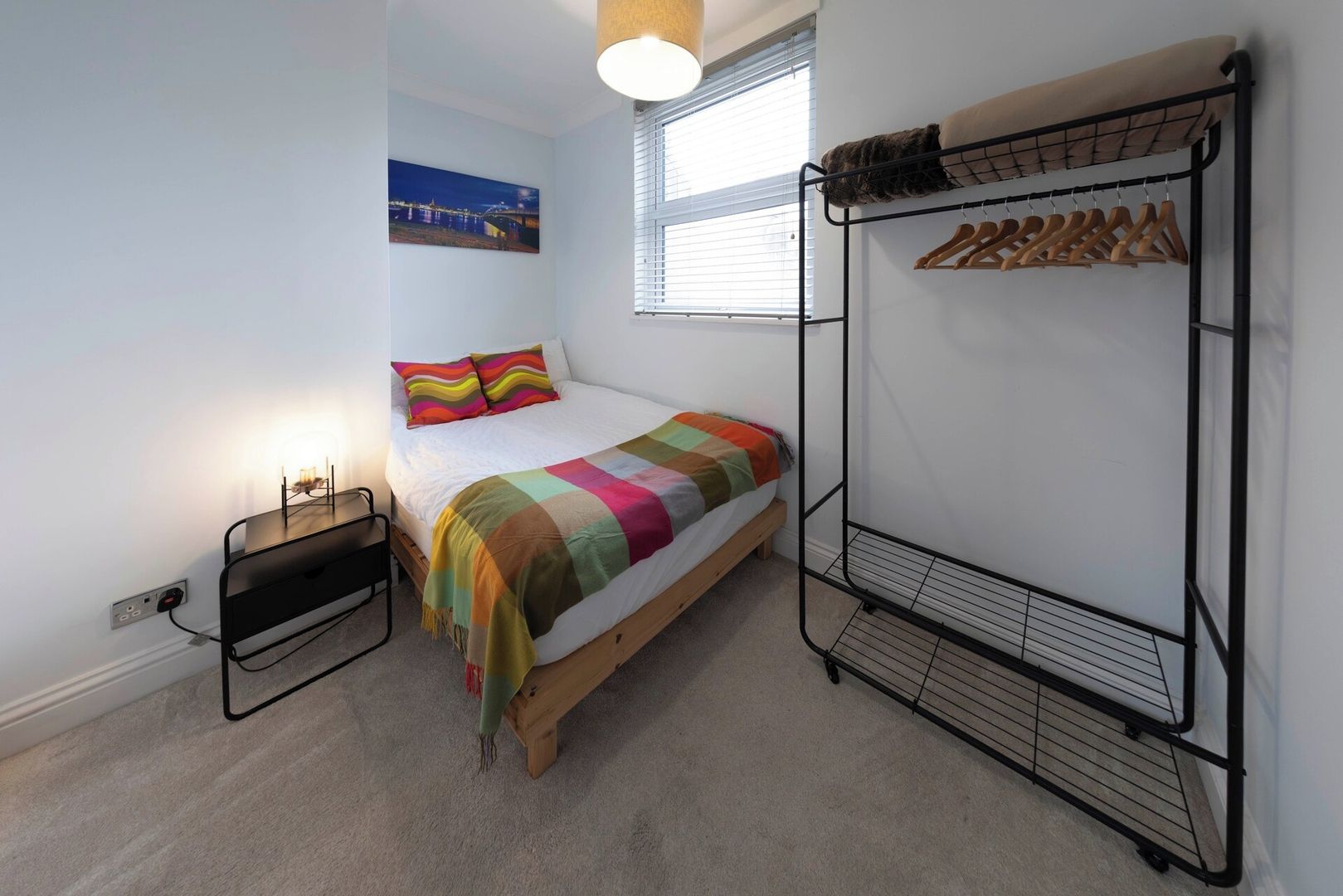 A bedroom with a bed, a bunk bed, a bedside table, a lamp, a window with blinds, and a wall-mounted coat rack.