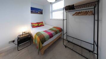Une chambre à coucher comprenant un lit, un lit superposé, une table de chevet, une lampe, une fenêtre avec des stores et un porte-manteau fixé au mur.
