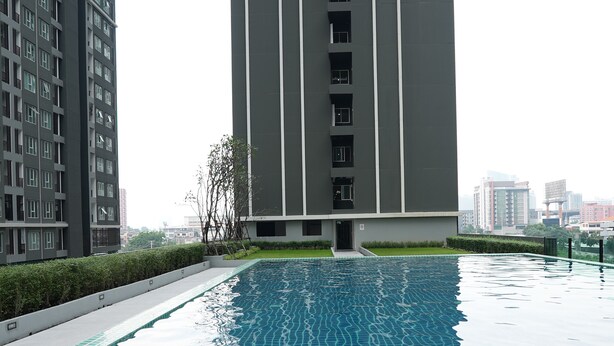 A swimming pool with a tiled edge and metal handrails, surrounded by a low hedge and a tall building with multiple windows. - A charming one-bedroom a condo near the BTS (Bangkok)