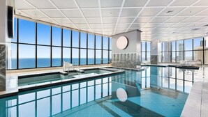 Una piscina interior moderna amb grans finestres que ofereixen vistes a l'oceà, un paviment de rajoles i un element decoratiu circular a la paret.
