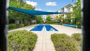 Ein rechteckiger Swimmingpool mit einer blauen Abdeckung, umgeben von gepflegten Büschen und einem gepflasterten Weg.