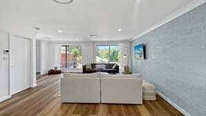 A modern living room with a large sectional sofa, a flat-screen TV mounted on a brick-patterned wall, and a large window providing a view of the outdoors. - Spacious 2-Bed Townhouse with BBQ (Surf Beach)