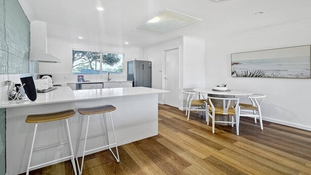 A modern kitchen with white countertops, wooden stools, and a dining area with white chairs. - Spacious 2-Bed Townhouse with BBQ (Surf Beach)