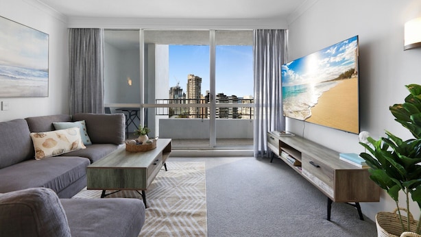 Un soggiorno moderno con un divano grigio, un tavolino da caffè e una TV su un supporto in legno. Una grande finestra offre una vista sulla città e sulla spiaggia.
