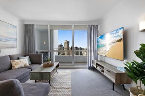 A modern living room with a grey sofa, a coffee table, and a TV on a wooden stand. A large window offers a view of a cityscape and a beach. - Ocean View 2-Bed with Pool, Tennis Court & BBQ (Surfers Paradise)