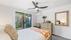 Une chambre à coucher comprenant un lit, un ventilateur de plafond, une commode, un miroir et une porte-fenêtre coulissante donnant sur un balcon agrémenté de plantes.