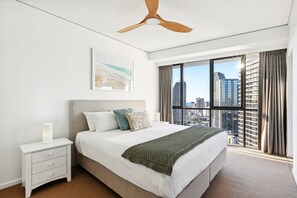 Ein modernes Hotelzimmer mit einem großen Bett, einem Deckenventilator, einem Nachttisch, einem Fenster mit Blick auf die Stadt und einem gerahmten Kunstwerk an der Wand.
