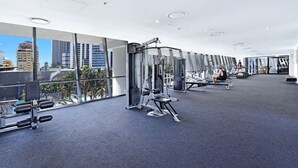 A modern gym with various exercise equipment, large windows offering city views, and a spacious layout. - Circle on Cavill Luxe 2-Bed with Views & Pool (Surfers Paradise)