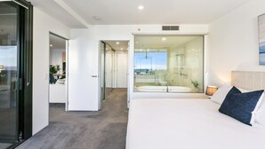 A modern hotel room with a large bed, a glass-enclosed bathroom, and a view of the cityscape. - Circle on Cavill Luxe 2-Bed with Views & Pool (Surfers Paradise)
