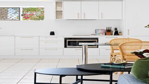 A modern kitchen with white cabinets, a microwave, and a dining area with a round table and wicker chair. - Chic 1-Bed Escape with Parking Bridgeman Downs (Bridgeman Downs)