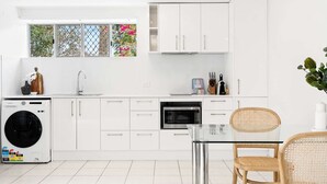 A modern kitchen with white cabinets, a washing machine, a microwave, and a dining area with a glass table and wicker chairs. - Chic 1-Bed Escape with Parking Bridgeman Downs (Bridgeman Downs)