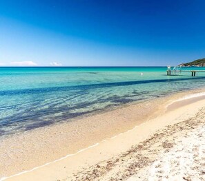 Ein Strand mit klarem türkisblauem Wasser, einem sandigen Ufer und einem Steg, der ins Meer führt.