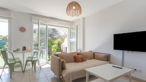 A modern living room with a beige sofa, a white table, and a TV mounted on the wall. There is a dining area with a table and chairs, and a large window providing a view of greenery outside. - Terrace apartment Puget-sur-Argens (Puget-sur-Argens)
