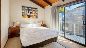Une chambre avec un grand lit, des poutres apparentes en bois au plafond, un tableau au mur, une table de chevet avec une lampe et une porte-fenêtre coulissante donnant sur l’extérieur.