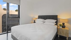 Moderni hotellihuone, jossa on suuri sänky, kaksi yöpöytää valaisimineen ja näkymä kaupunkiin lattiasta kattoon ulottuvien ikkunoiden kautta.