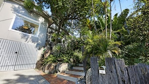 Um imóvel residencial com fachada branca, um caminho de tijolos que leva a um portão de madeira e uma variedade de plantas tropicais.