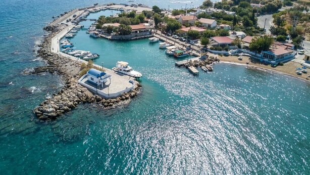 Vista aérea de un pueblo costero con un puerto deportivo, costa rocosa y edificios con tejados rojos.