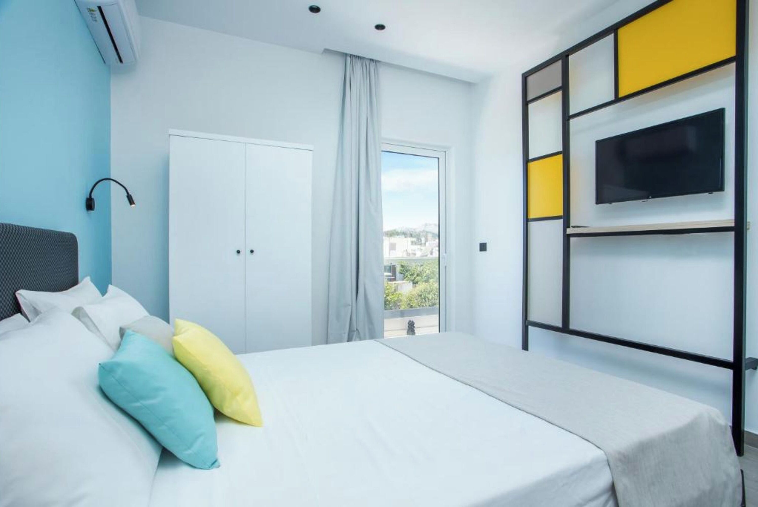 Habitación de hotel con cama, televisor montado en la pared, ventana con cortinas y vistas a la ciudad.