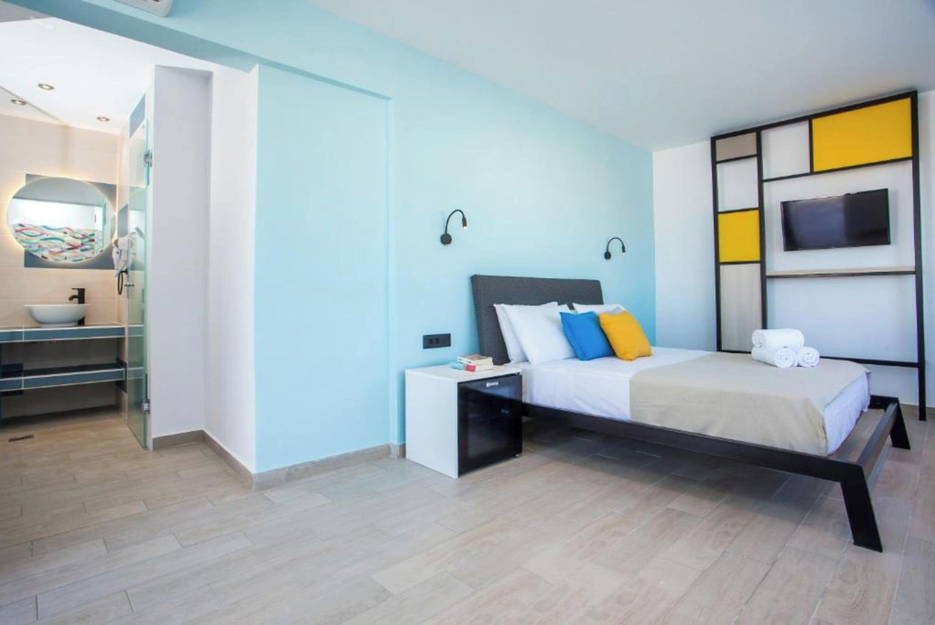 A modern bedroom with a bed, bedside tables, a TV, and a bathroom visible in the background.