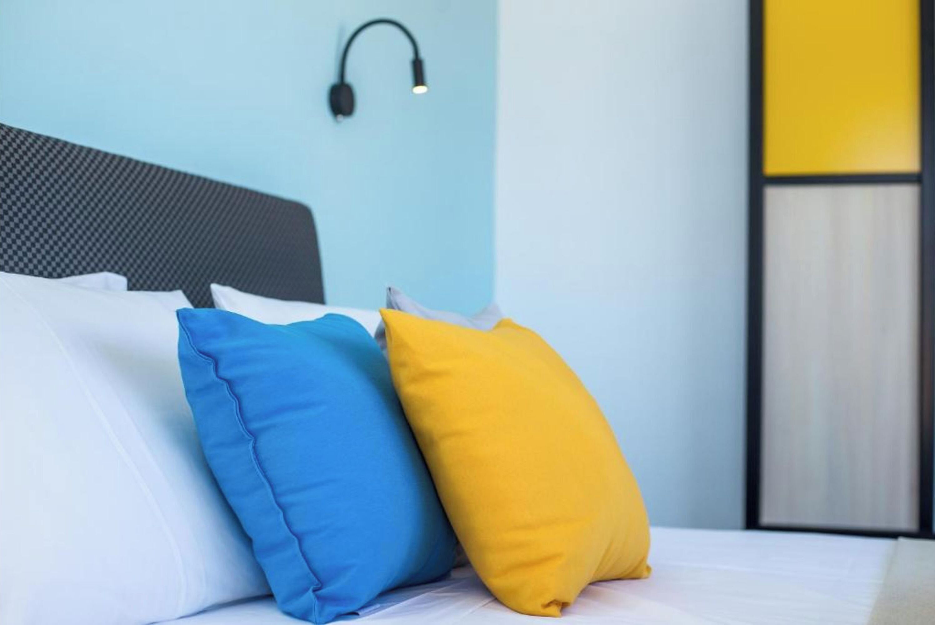 A bed with white bedding and two pillows, one blue and one yellow, in a room with a light blue wall and a black headboard.